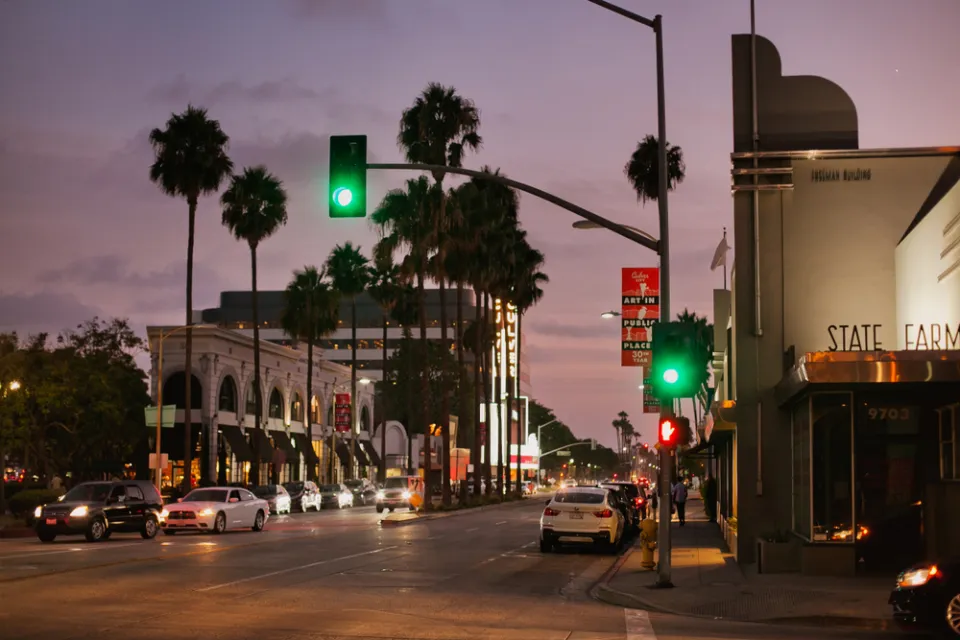 View of Culver City street with stoplights, white cars, and palm trees sillhouetted against a purple sunset sky
